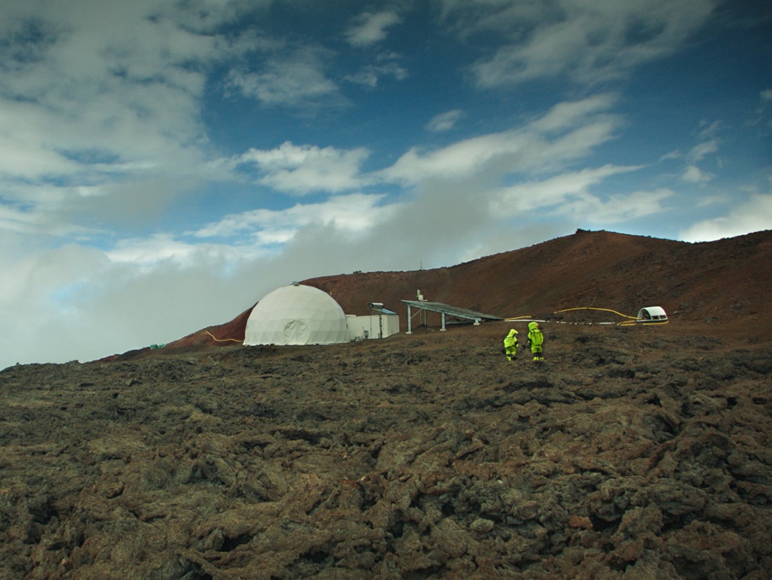 Marslandschaft auf Hawaii, zwei Personen in neonfarbener Schutzkleidung, ein weißes, Iglu-ähnliches Zelt und Solarpaneele.