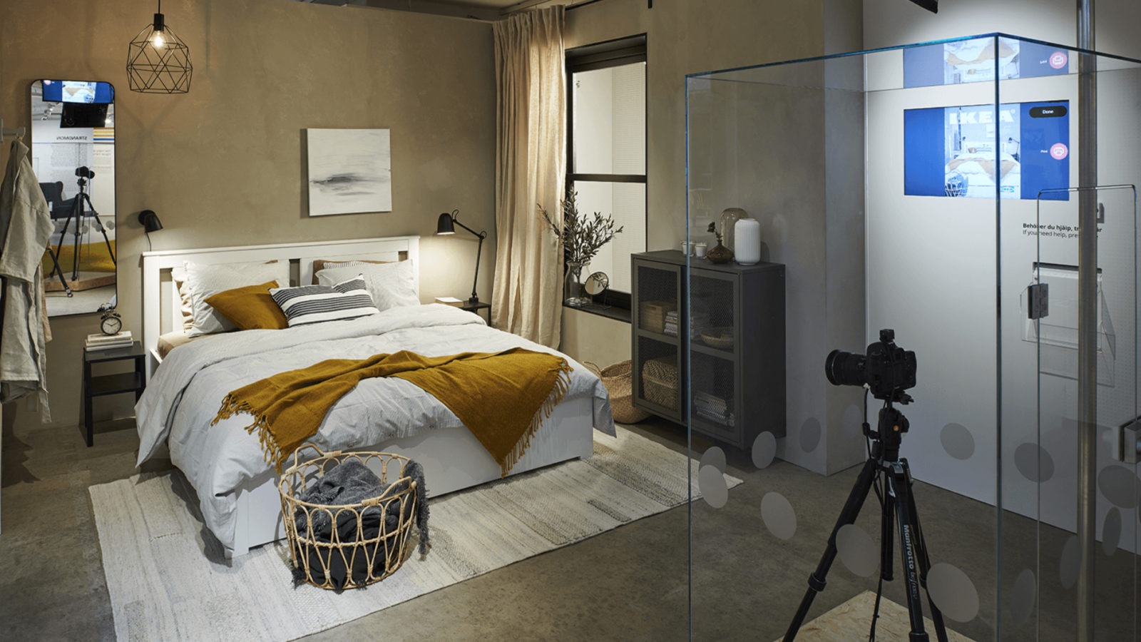 Ein gemütliches Schlafzimmer mit einem gemachten Doppelbett, einem Teppich, Kissen und Lampen, und einem großen Glaskasten, indem sich eine Kamera auf einem Stativ befindet.