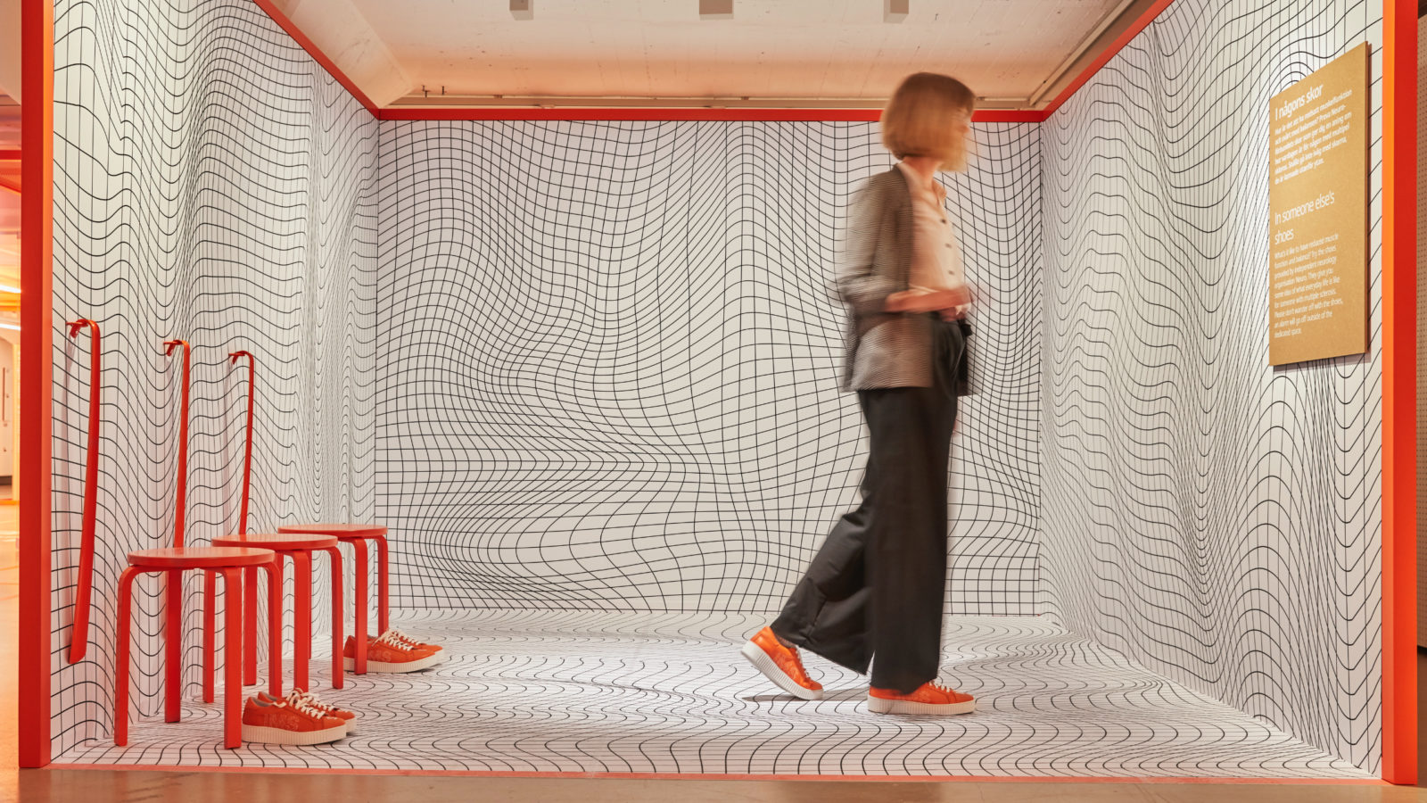 A woman inside a space covered with black and white geometrical patterns, three bright orange stools, shoes and shoe horns.