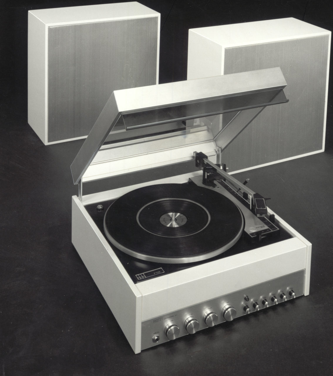 Ein schwarz-weißer RENN Plattenspieler aus den 1970er-Jahren mit einem durchsichtigen, geöffneten Plattendeckel und zwei weißen Lautsprechern im Hintergrund.