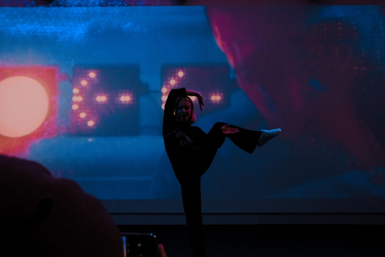 Eine Frau, die beim Tanzen in einem großen, schwach beleuchteten Raum vor einem blauen Hintergrund mit pfeilförmigen Lampen ihr Bein streckt.
