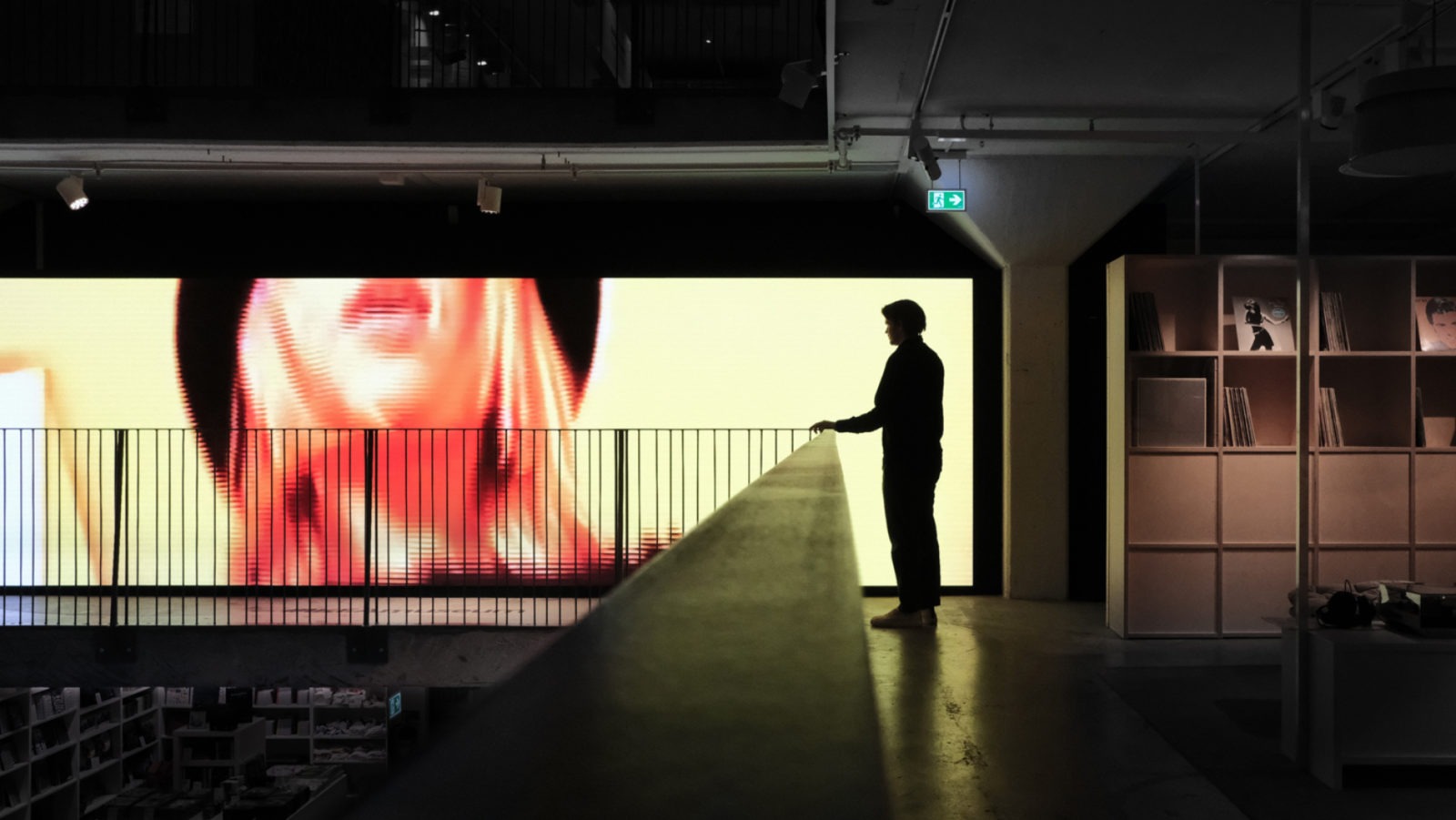 The silhouette of a person standing inside IKEA Museum, looking at a large screen showing a woman enjoying music.
