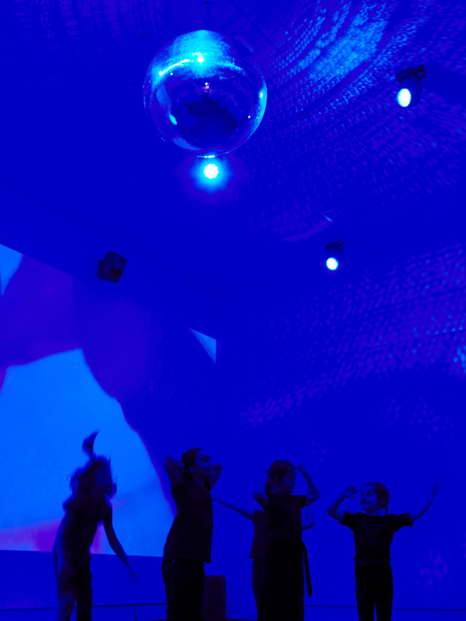 Menschen unterschiedlichen Alters tanzen in einem großen Raum mit blauem Licht und einer Discokugel, die von der Decke hängt.