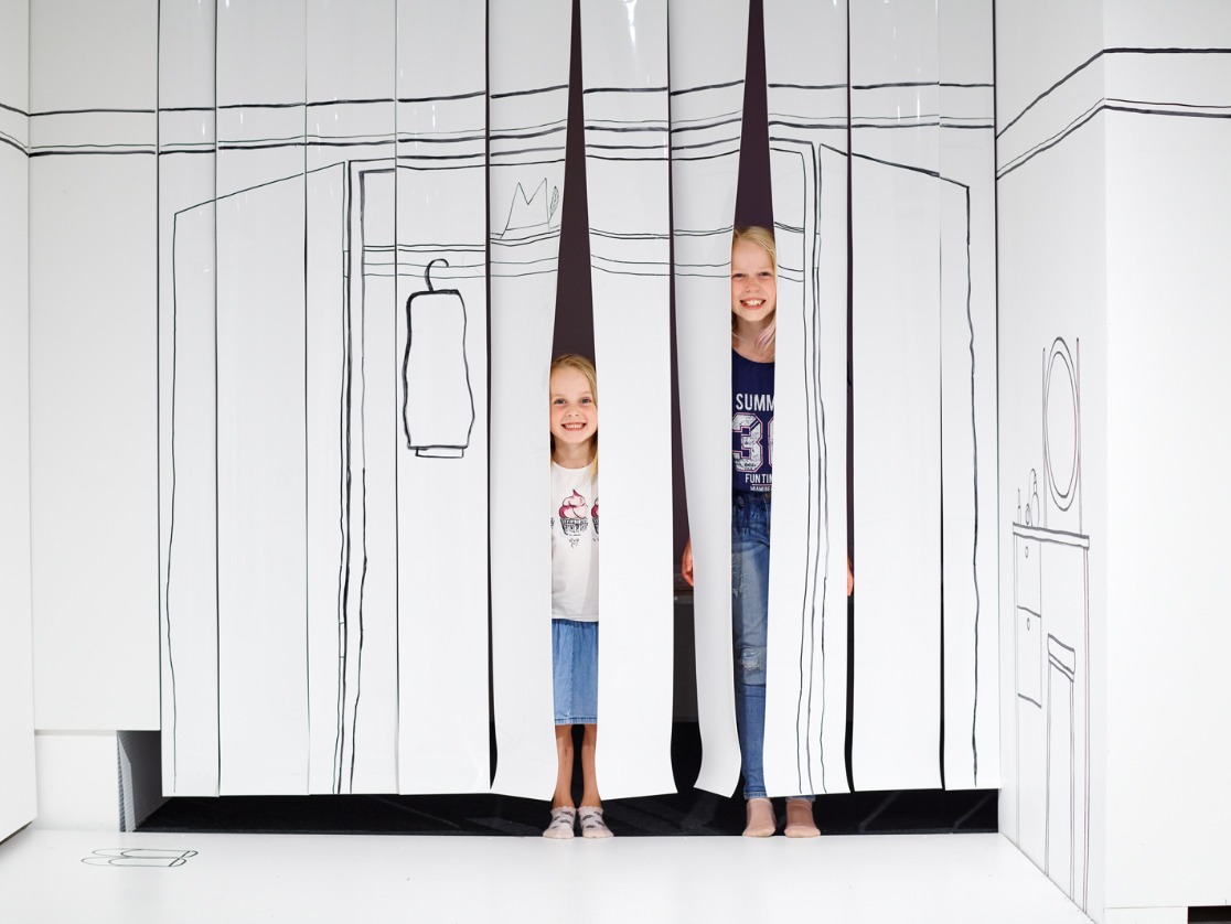 A white space with furniture drawings in black and white, and two smiling girls looking through curtain sleeves.