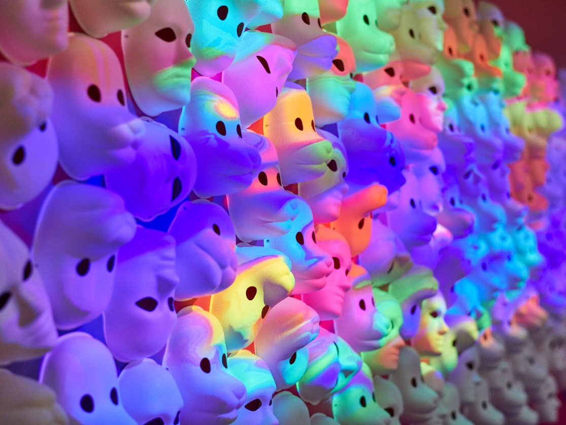 Face masks for play covering a large wall completely, bathed in multicoloured, fluid light.