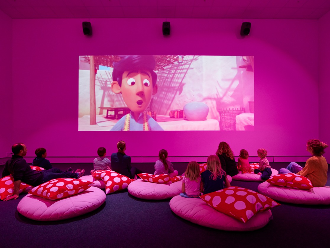 A room bathed in pink light with children and some adults sitting on flat pouffes on the floor, watching an animated film.