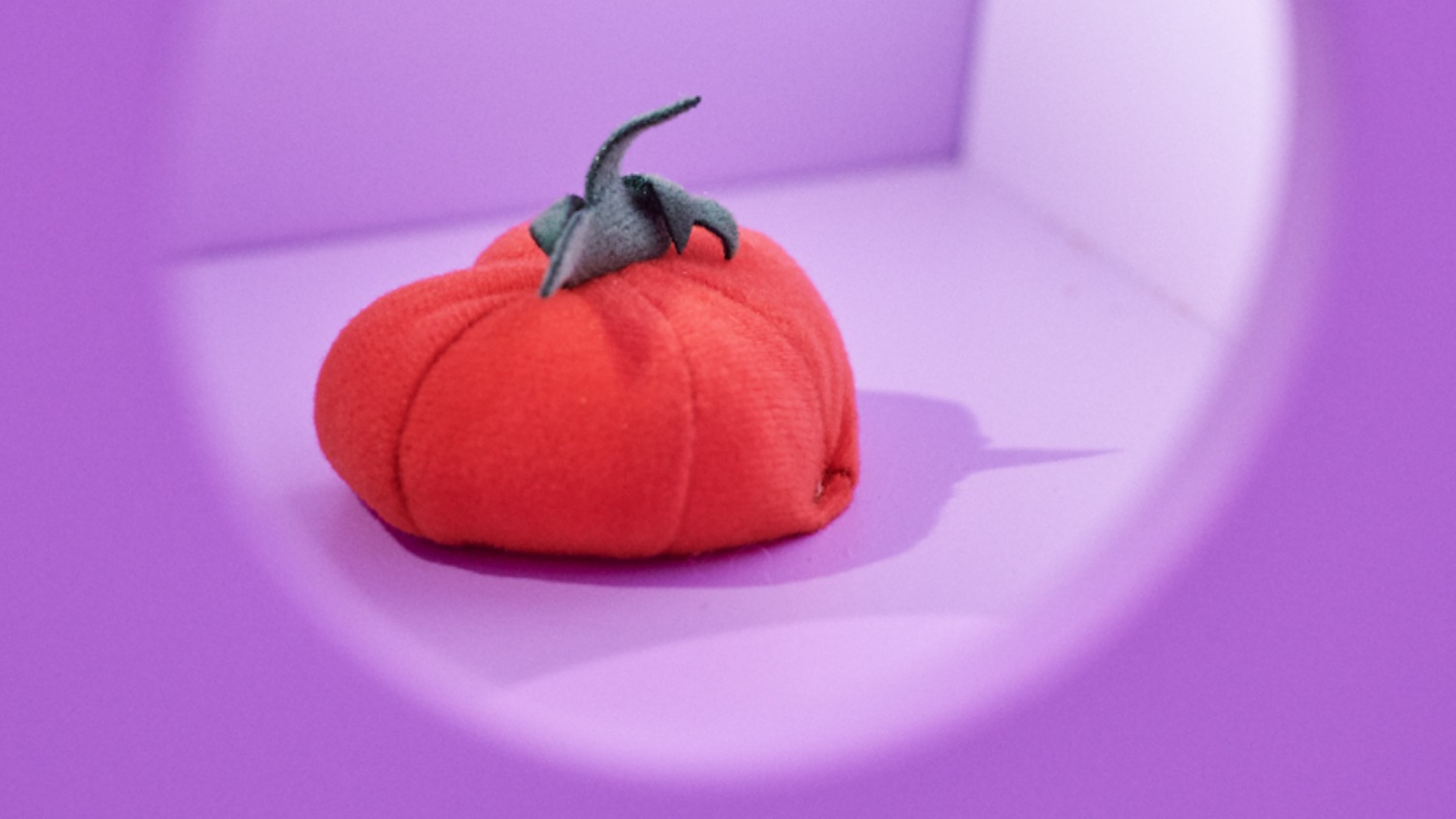 Durch ein violettes Guckloch ist eine rote Tomate aus Stoff zu sehen.