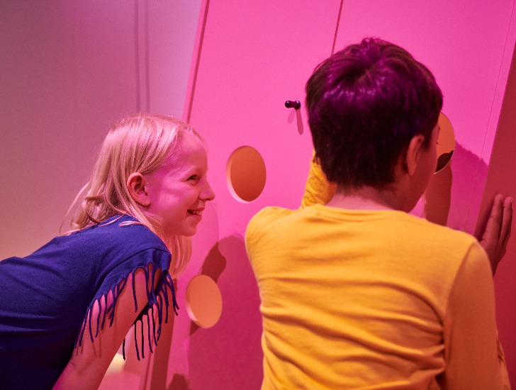 En flicka och en pojke badar i rosa ljus och kikar in genom hålen i en tittut-garderob.