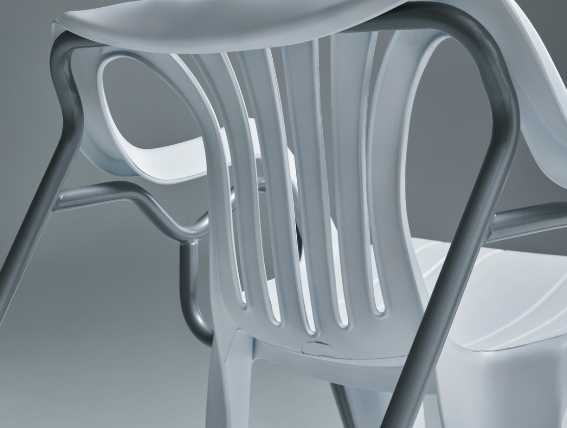 A detail of a plastic chair with armrests, intertwined with a metal shape that is partly supporting the plastic chair.