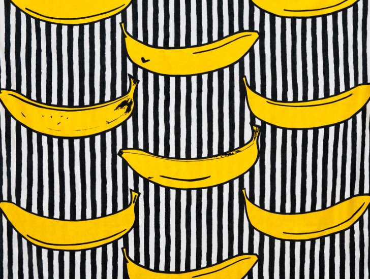 Der Stoff RANDIG BANAN mit einem plakativ-naiven Muster von gelben Bananen vor einem schwarz-weiß gestreiften Hintergrund.