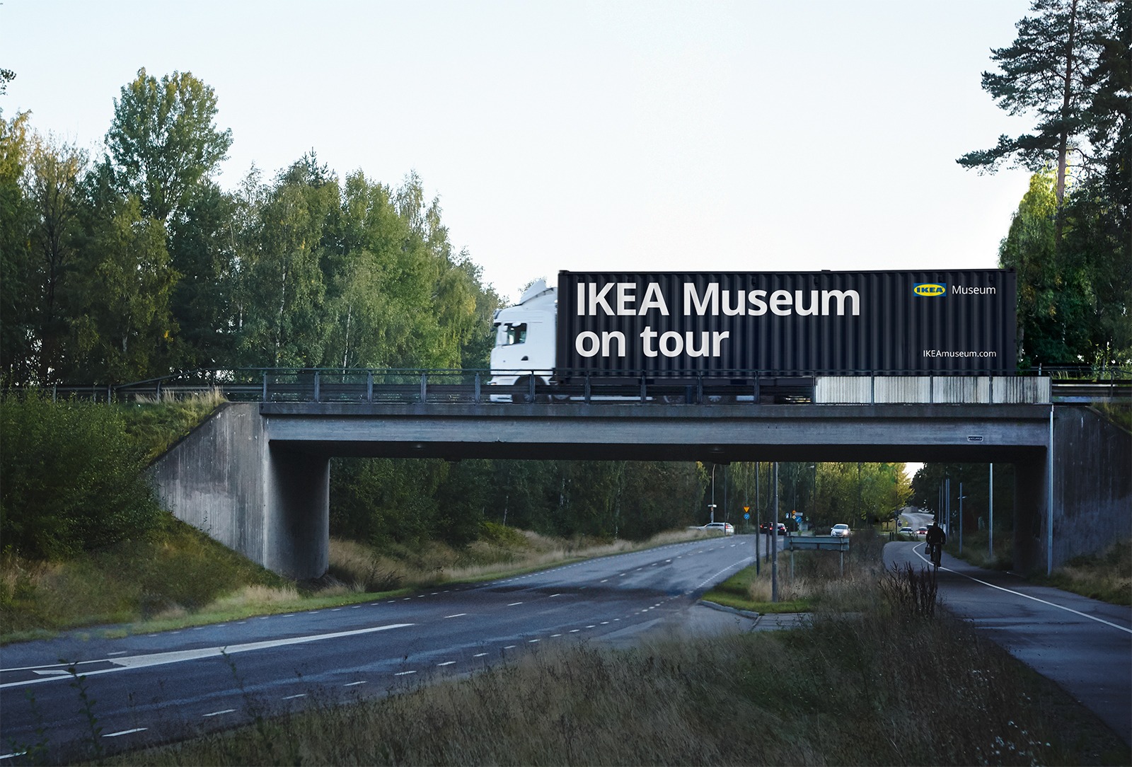 Trees, a road and a road bridge with a truck loaded with a container marked “IKEA Museum on tour” driving by.