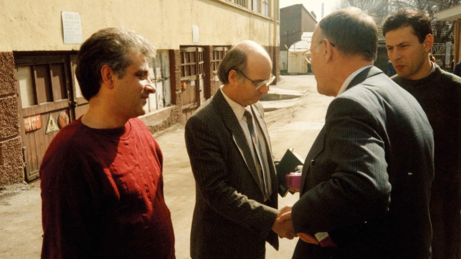 Ingvar Kamprad shakes the hand of a man in a suit, in an industrial area, outdoors. Two men watch the interaction.