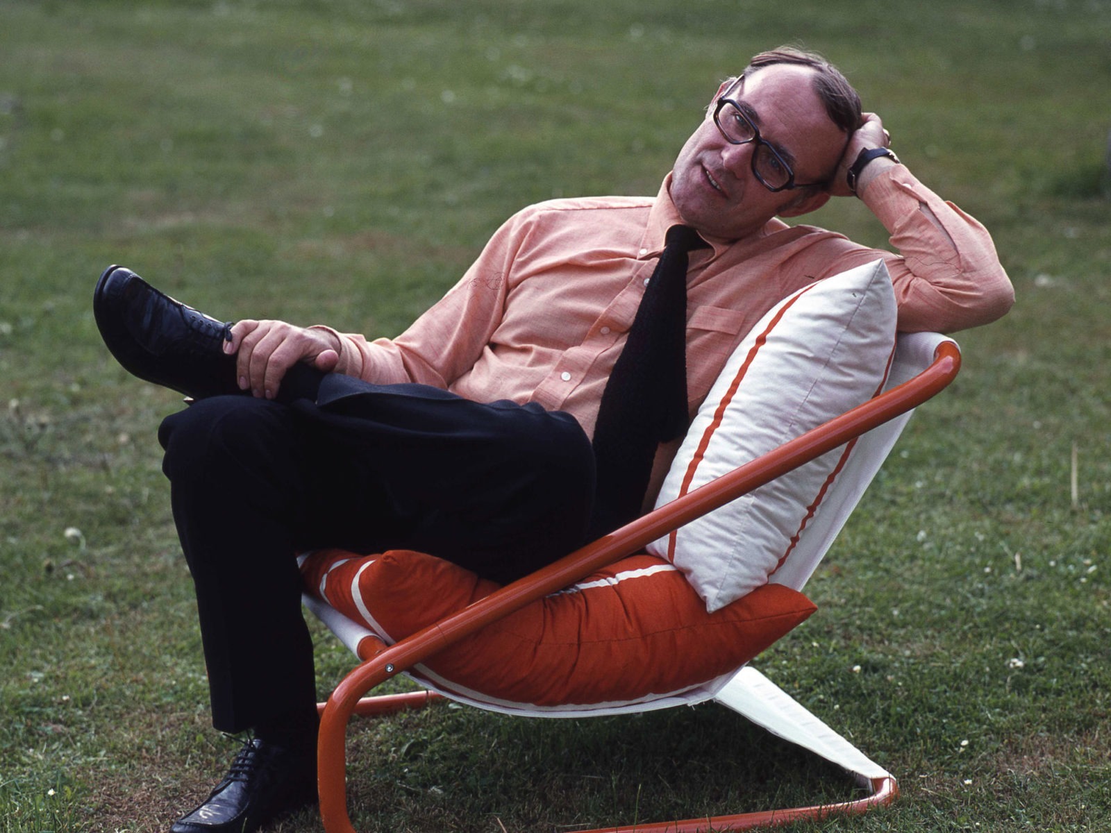 Ingvar Kamprad in pink shirt and black tie, relaxing in a 1970s style red and white chair on a lawn.