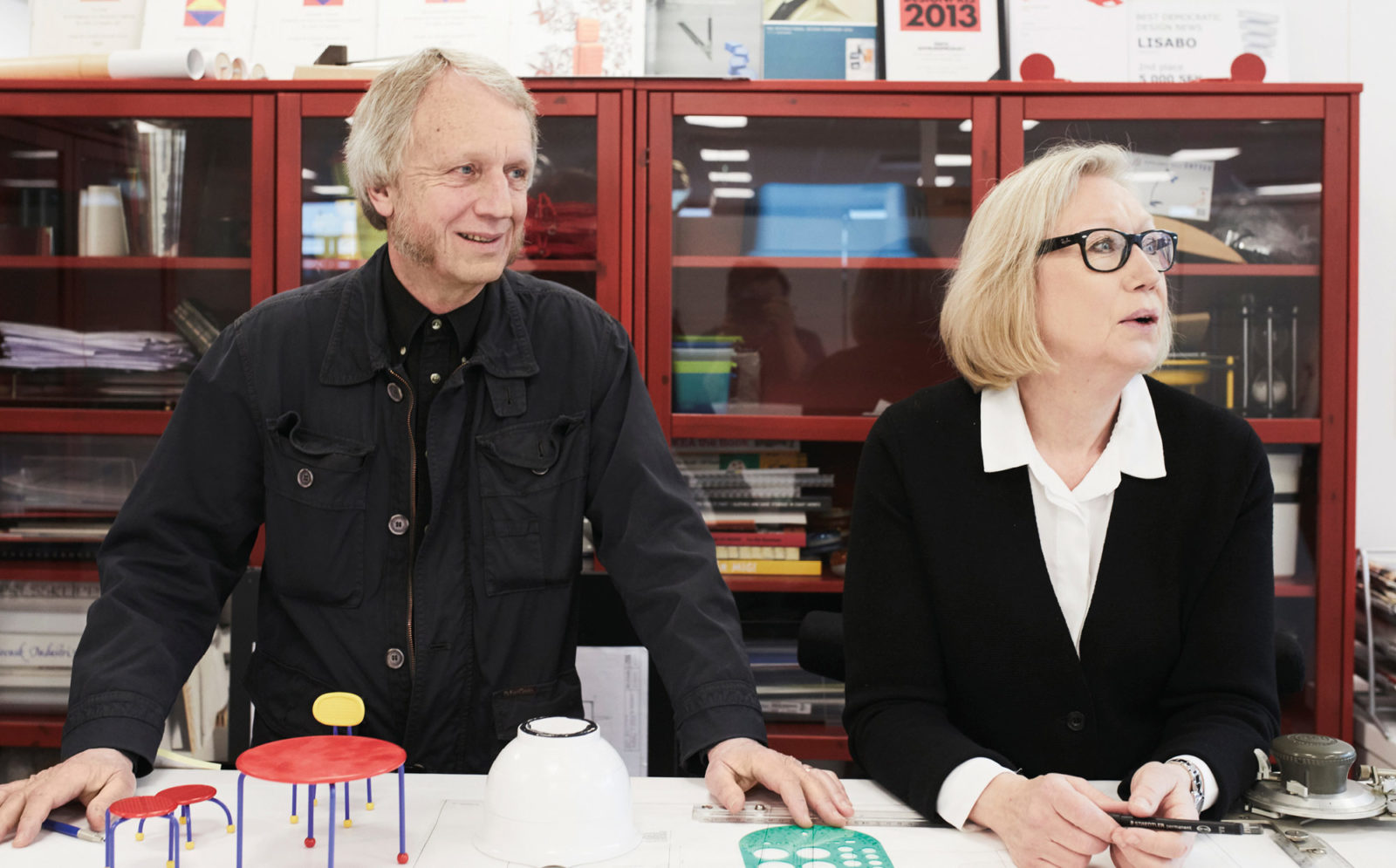 Middle-aged man and woman, blond, greying hair and black clothes, standing at a drawing table with prototypes and rulers.