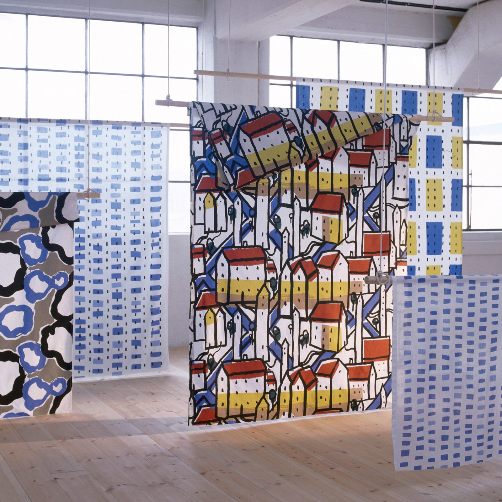 Fem textilier hänger från taket i en utställning, djärva mönster i en mängd olika färger.
