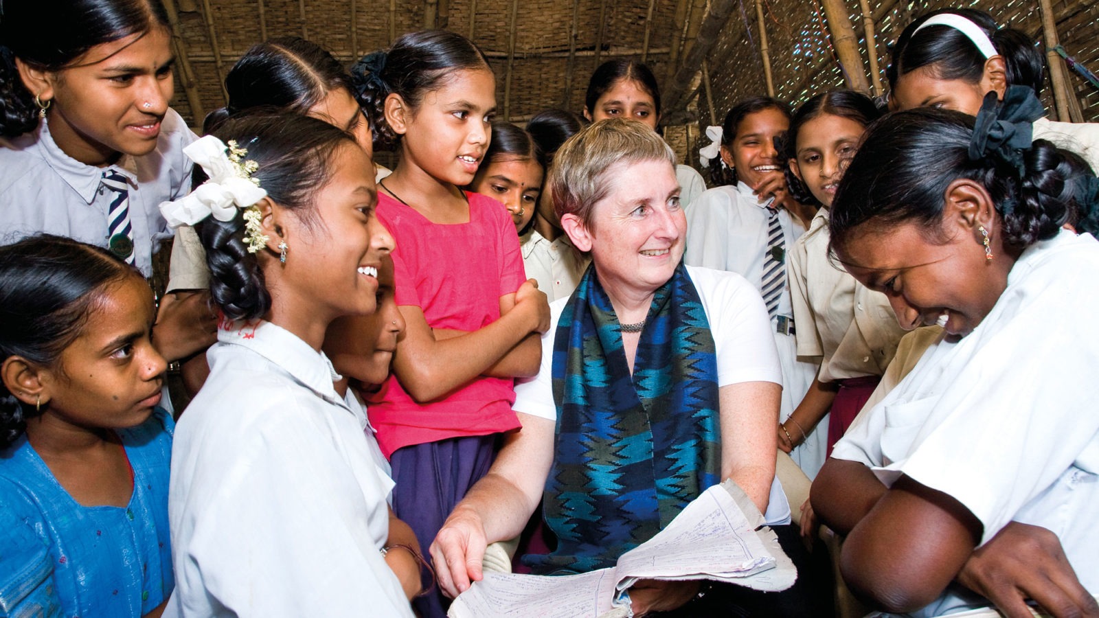 Marianne Barner with blond short hair and blue scarf, surrounded by laughing Indian school girls.