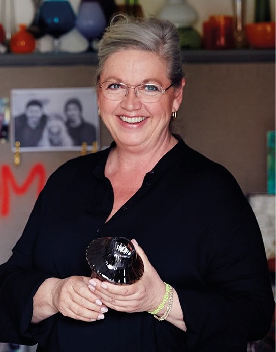 Smiling woman with short blonde hair and gold rimmed glasses, black shirt, yellow bracelet, holding ceramic candlestick.
