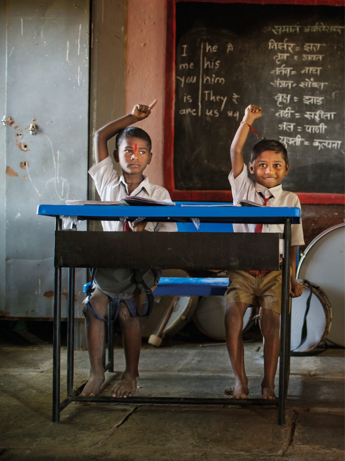 Two boys, all dressed in school uniforms sit with their hands raised in worn-down classroom. One boy smiles.