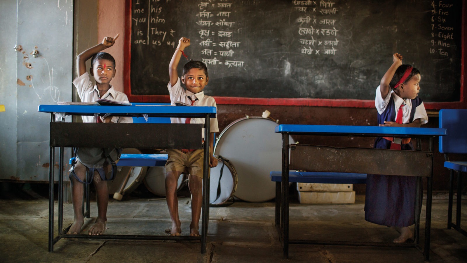 Two boys, one girl, all dressed in school uniforms sit with their hands raised in worn-down classroom. One boy smiles.