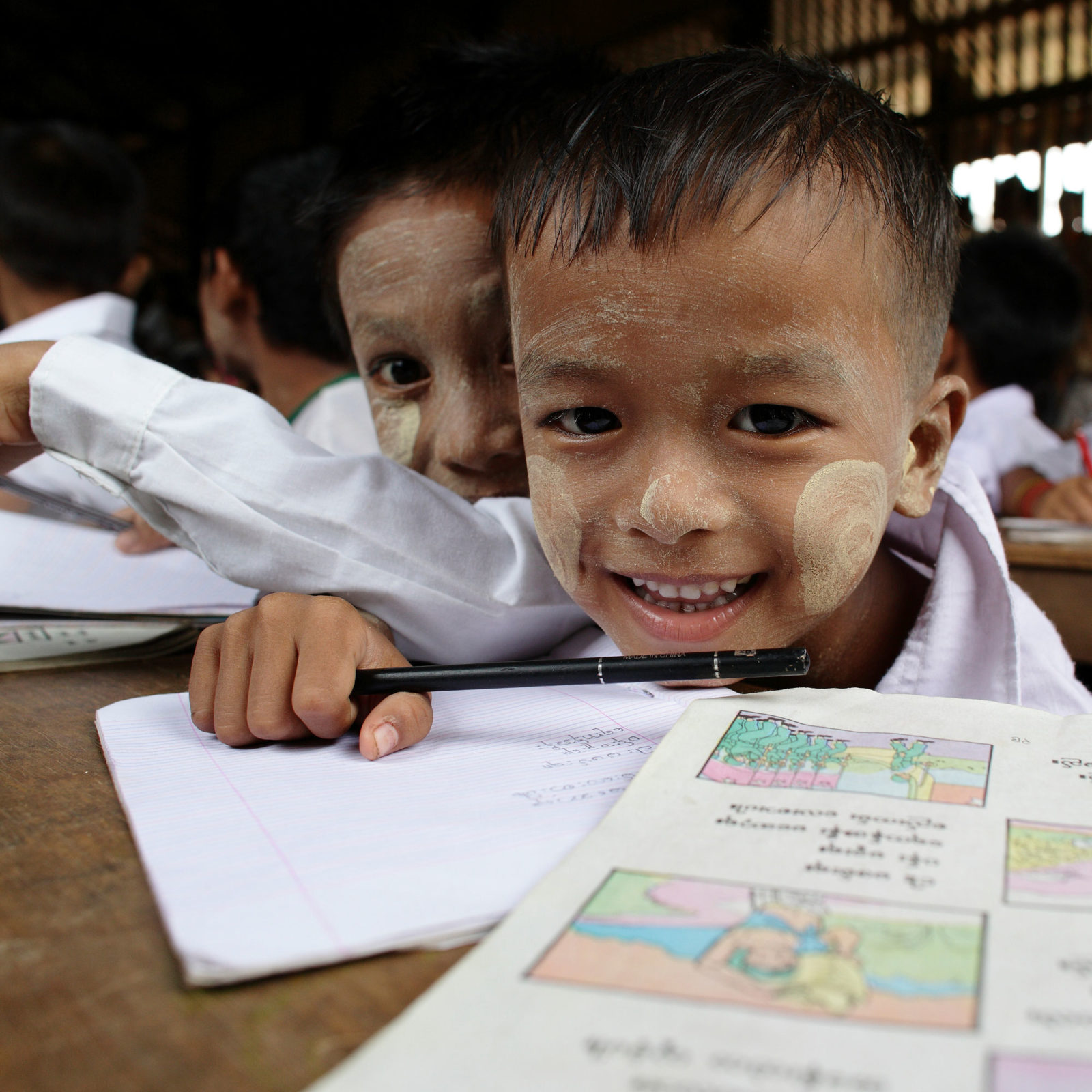 Close-up of smiling boy with, in Myanmar, traditional white markings on face, sitting in classroom, pen in hand.