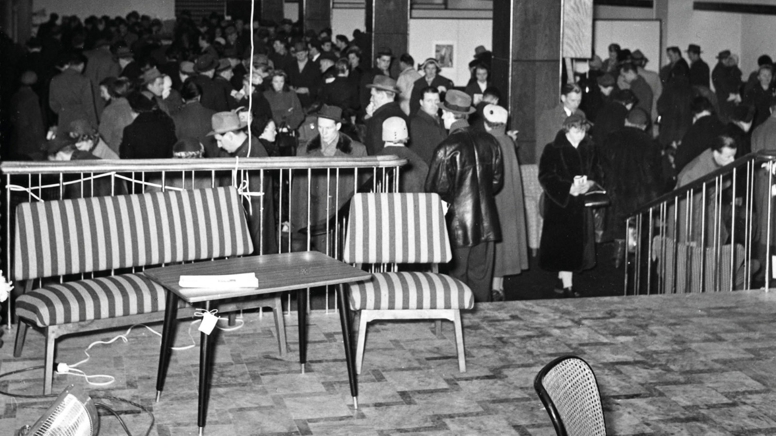 Randig soffa, träbord i 1950-talsstil uppställt i mässlokal. I bakgrunden syns myller av folk.
