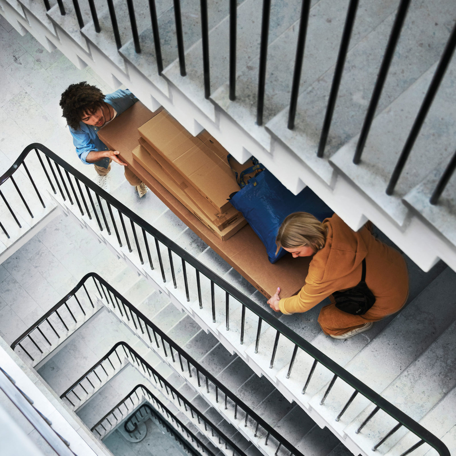 En ung man och kvinna bär tillsammans stora och till synes tunga platta paket från IKEA uppför trappor i ett trapphus.