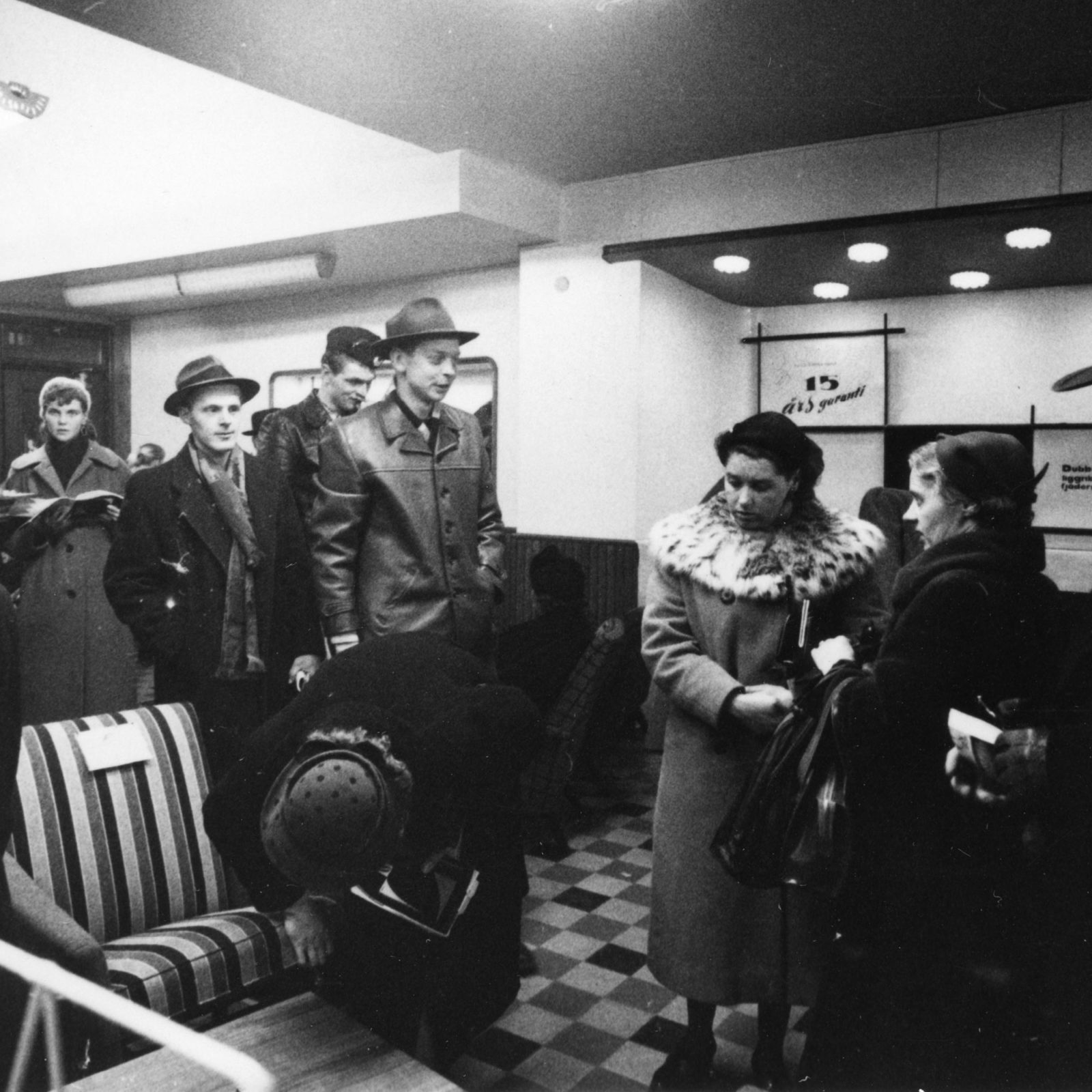Visitors crammed in at a 1950s IKEA furniture show, looking at posters and stuffed armchairs.
