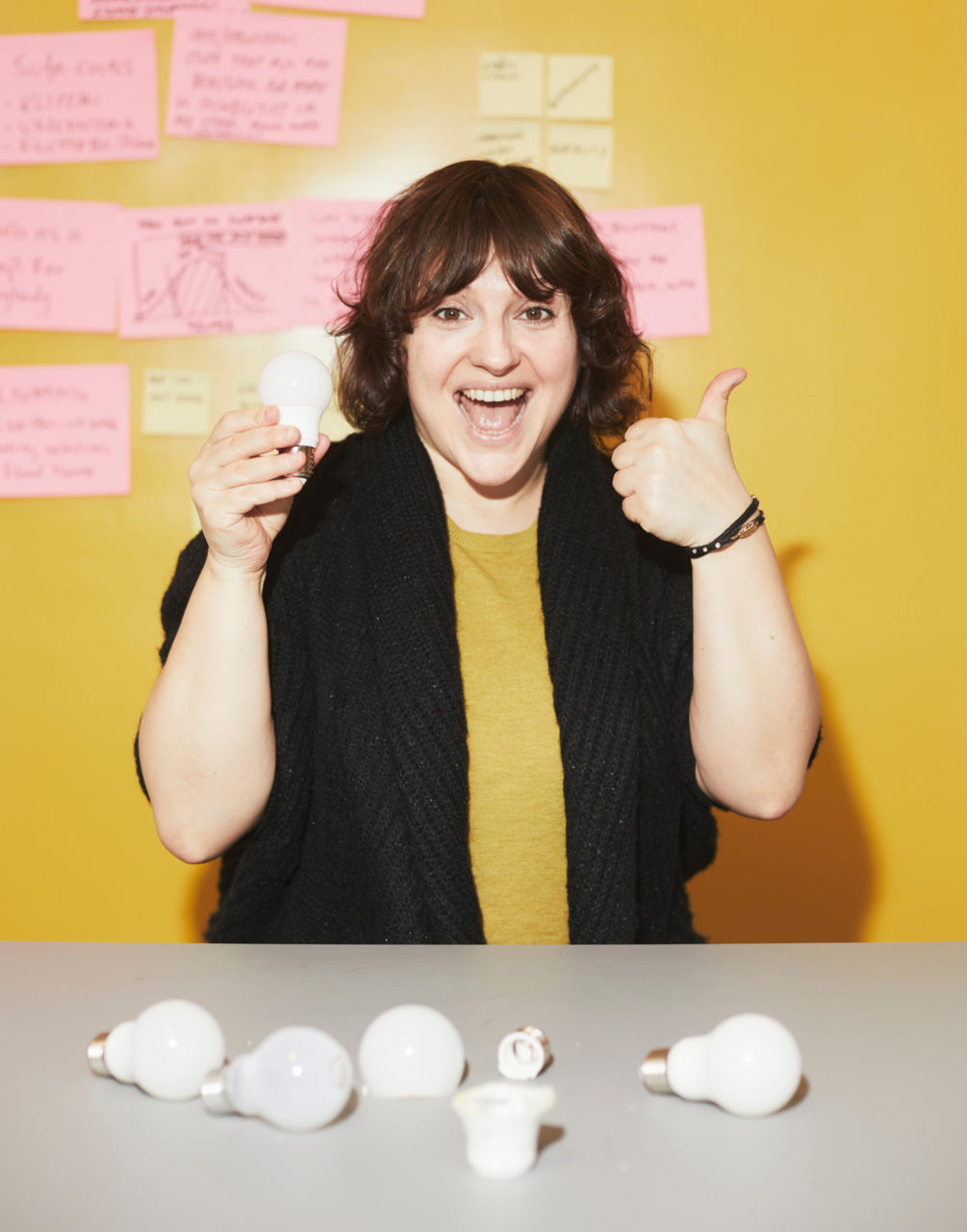 Paulina Pajak, smiling, sits at table scattered with LED bulb parts, holding a LED bulb and making a thumbs-up.