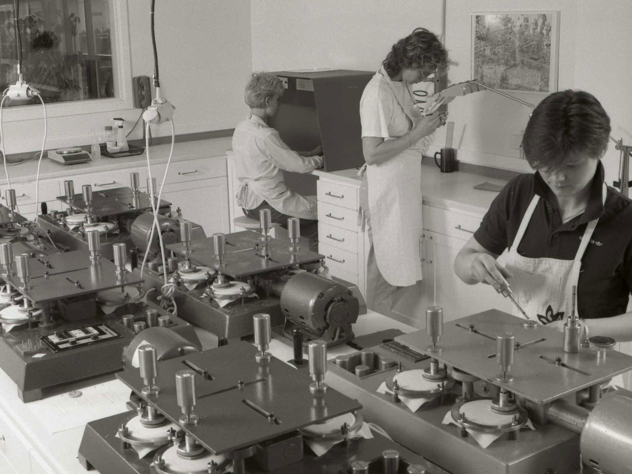 Three women checking things in a test laboratory, in machines or under strong lamps and magnifying glasses.