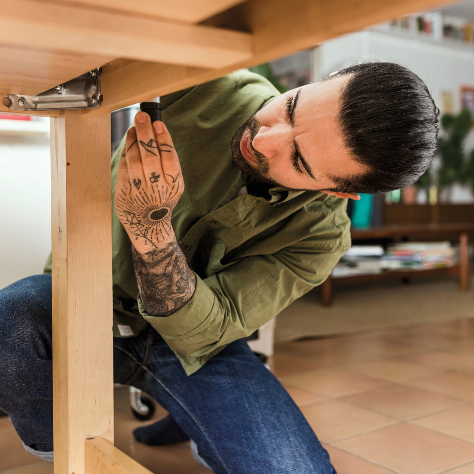 Young dark-haired man with beard and tattoos, wearing jeans and a green shirt, squats while assembling a table.