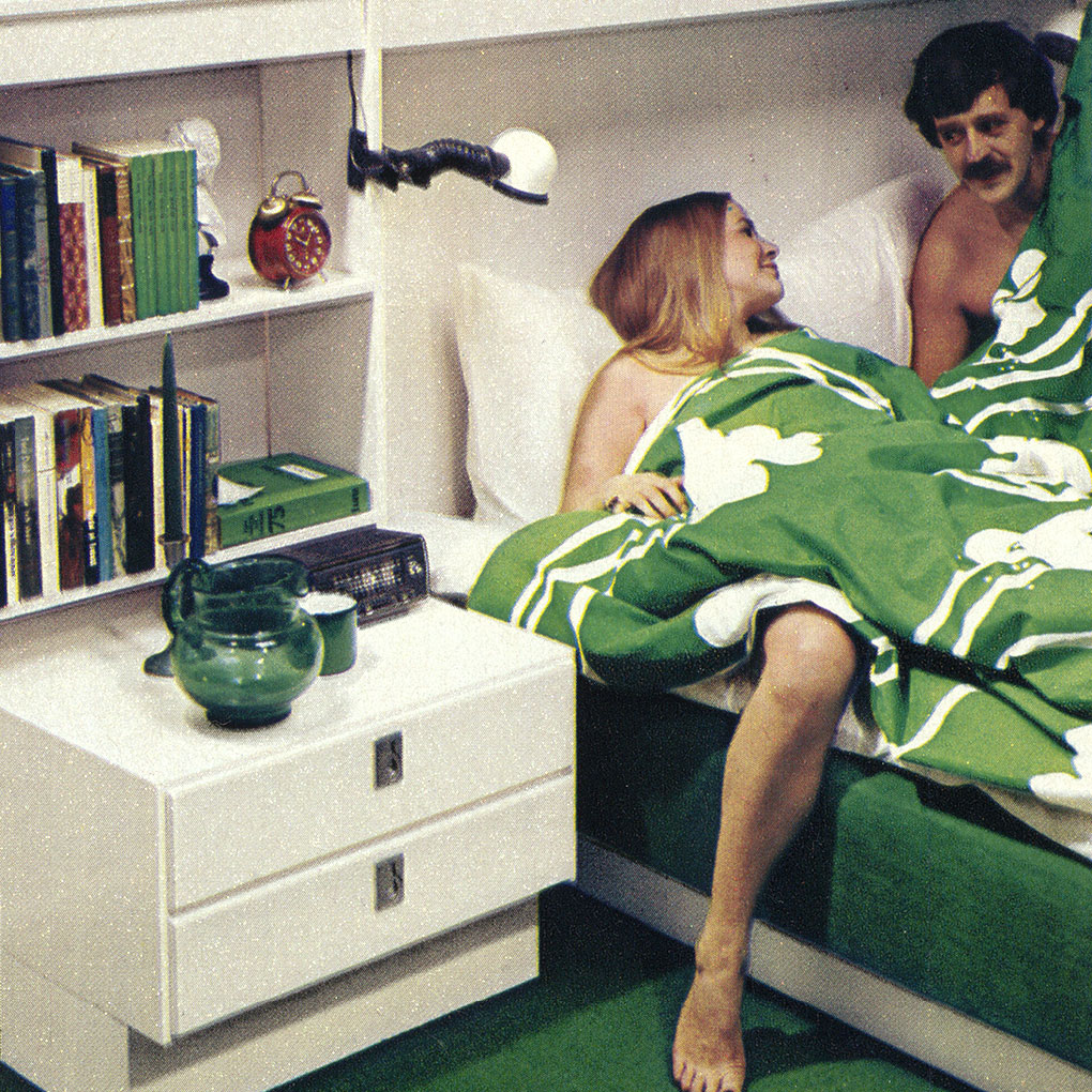 A woman and a man in bed under a white patterned green duvet, in a bedroom also completely white and green.