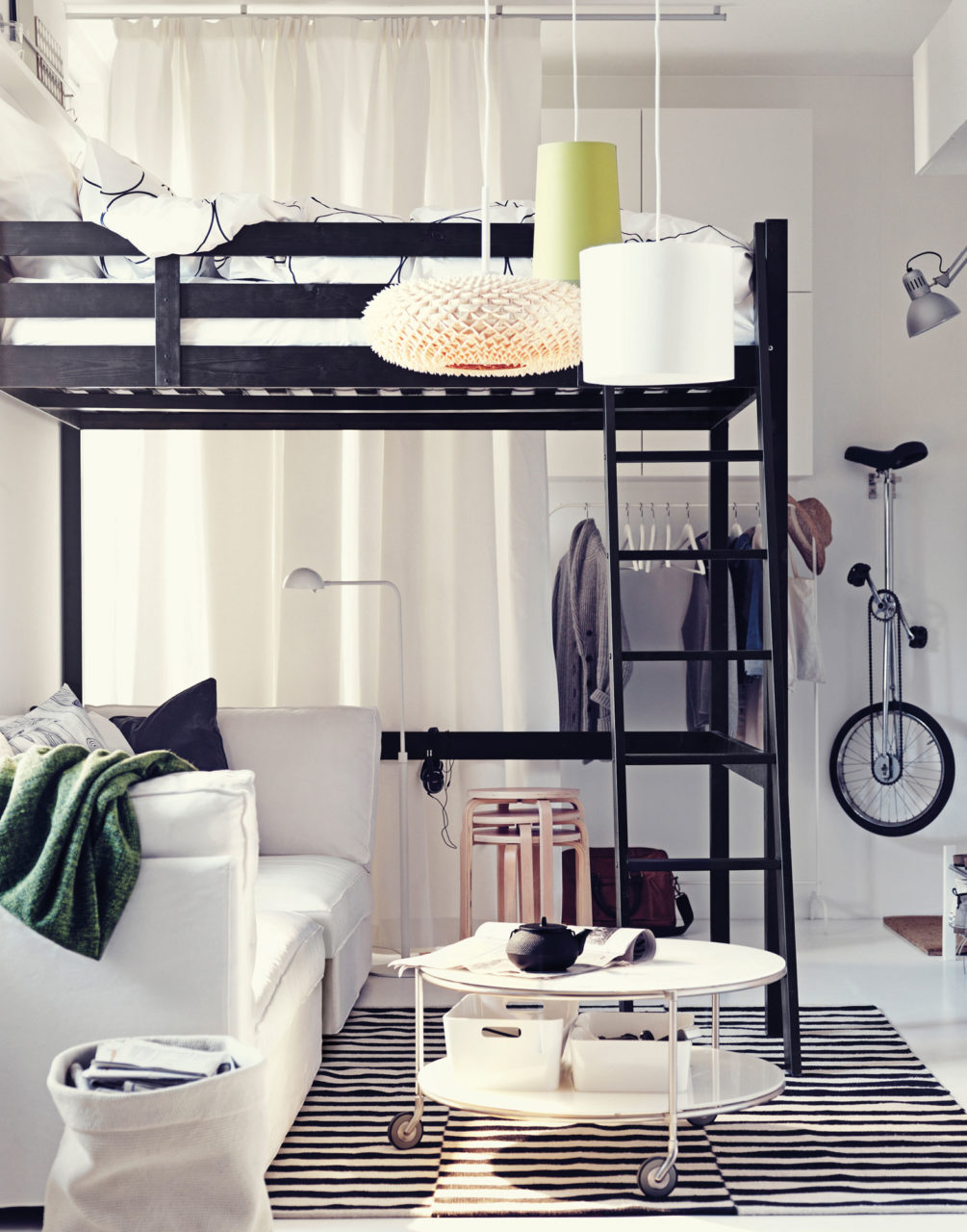 A light room with a white sofa standing under a high, black loft bed in a light style. Clothes and a unicycle are hanging on the wall.