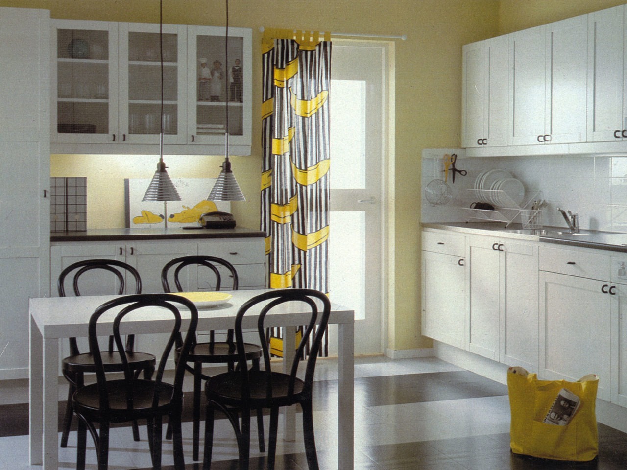 A kitchen in white apart from pin-back chairs at a dining table and details in black, and a banana-patterned curtain at the kitchen entrance.