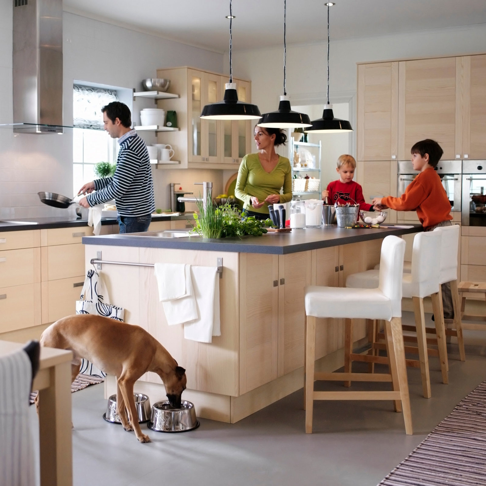 A man, a woman, two children and a dog spread around a large kitchen island with bar stools along one side. The kitchen is in light wood.