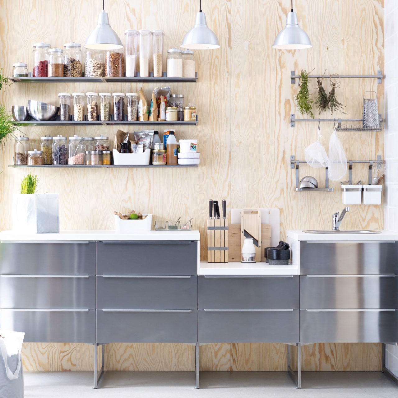 A modular kitchen with drawer fronts in brushed metal against a wall of untreated plywood. Dry goods in rows on narrow wall shelves.