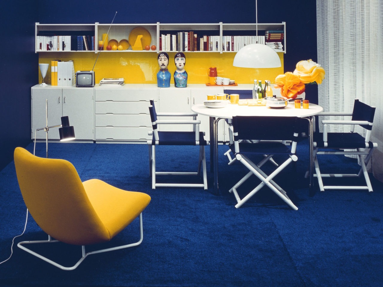 A completely blue, white and yellow room A long, white piece of storage furniture, director’s chairs around a dining table, a banana-like easy chair, blue rug.