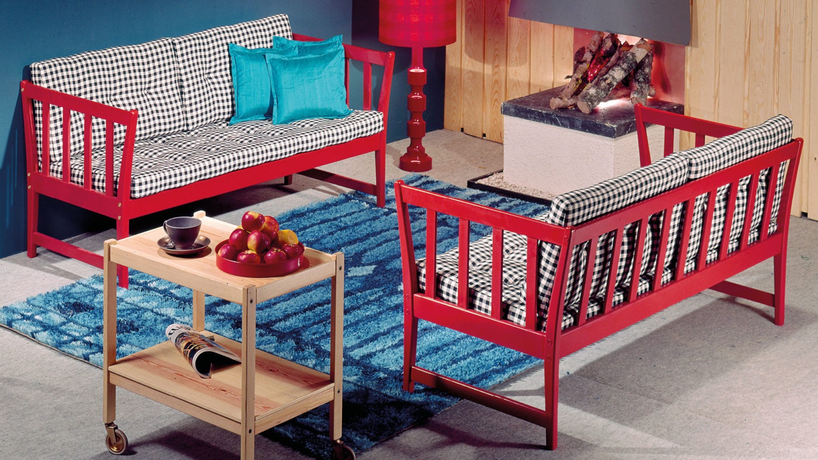 Sofás de estilo rústico con armazón rojo y cojines de cuadros. Carrito de madera color claro y alfombra azul.
