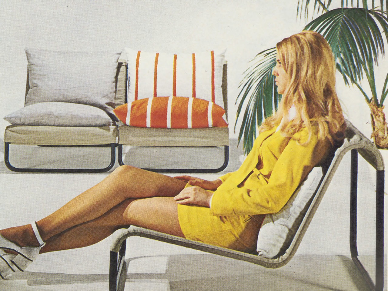 A woman in a low KRUMELUR armchair, which is textile over a metal frame. In profile, the chair forms an asymmetric M shape.