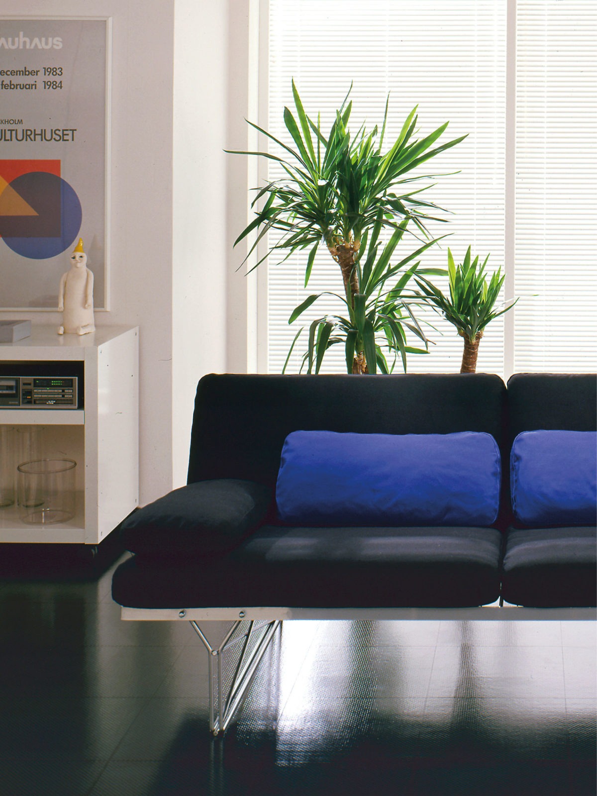 A black MOMENT sofa and a multicoloured MOMENT shelf with thin metal frames, in a room with a shiny black floor.
