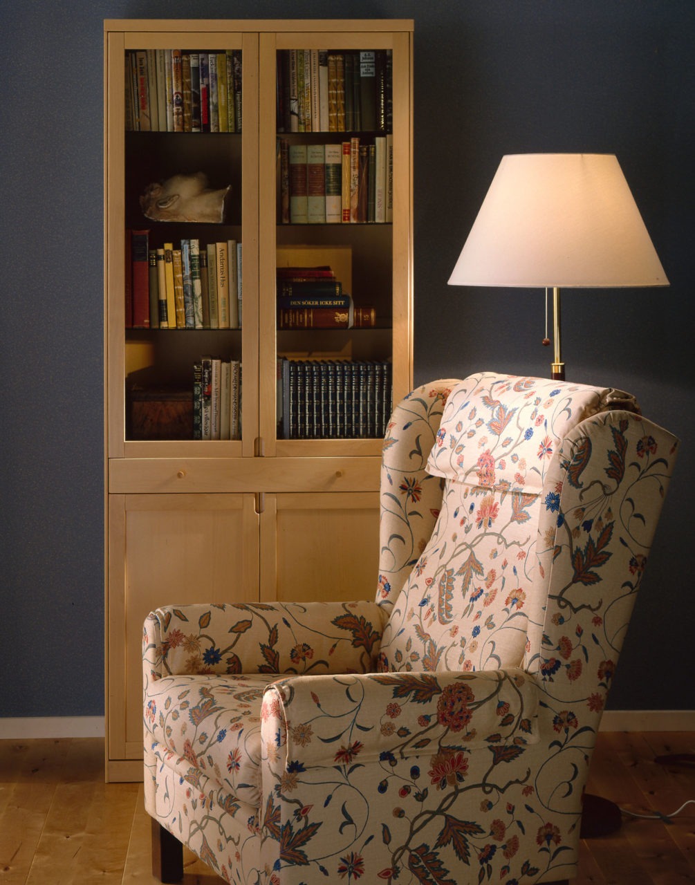 A light STOCKHOLM armchair with a plant pattern in bright colours stands next to a floor lamp and a tall glass-door cabinet.