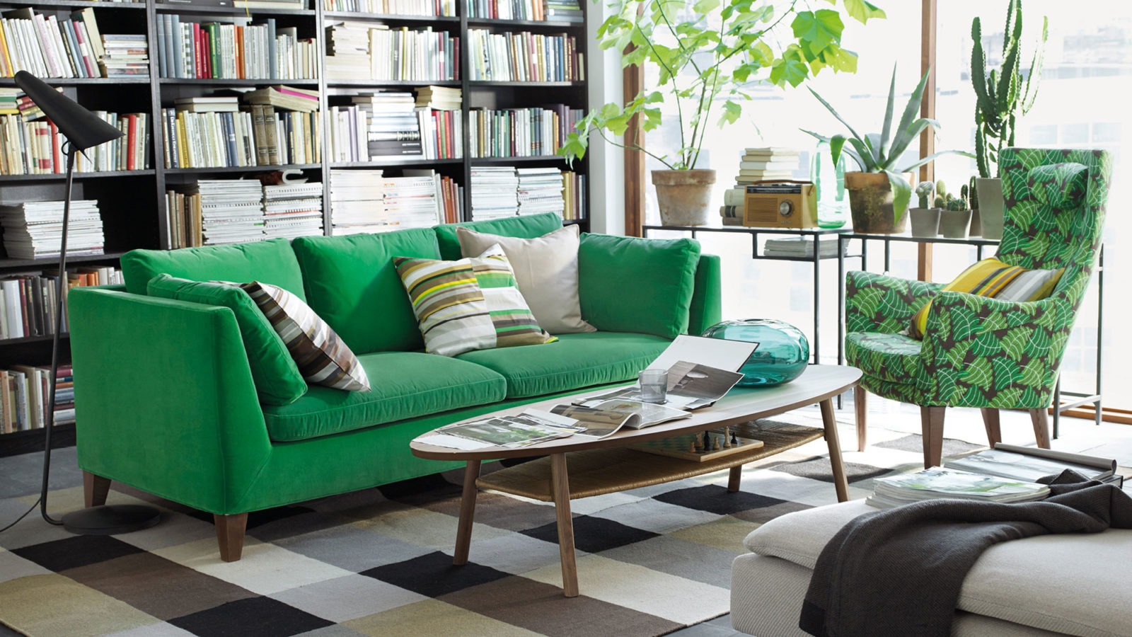 A room with a green sofa and a green-patterned armchair at an oval coffee table. Green plants in a large sunlit window.