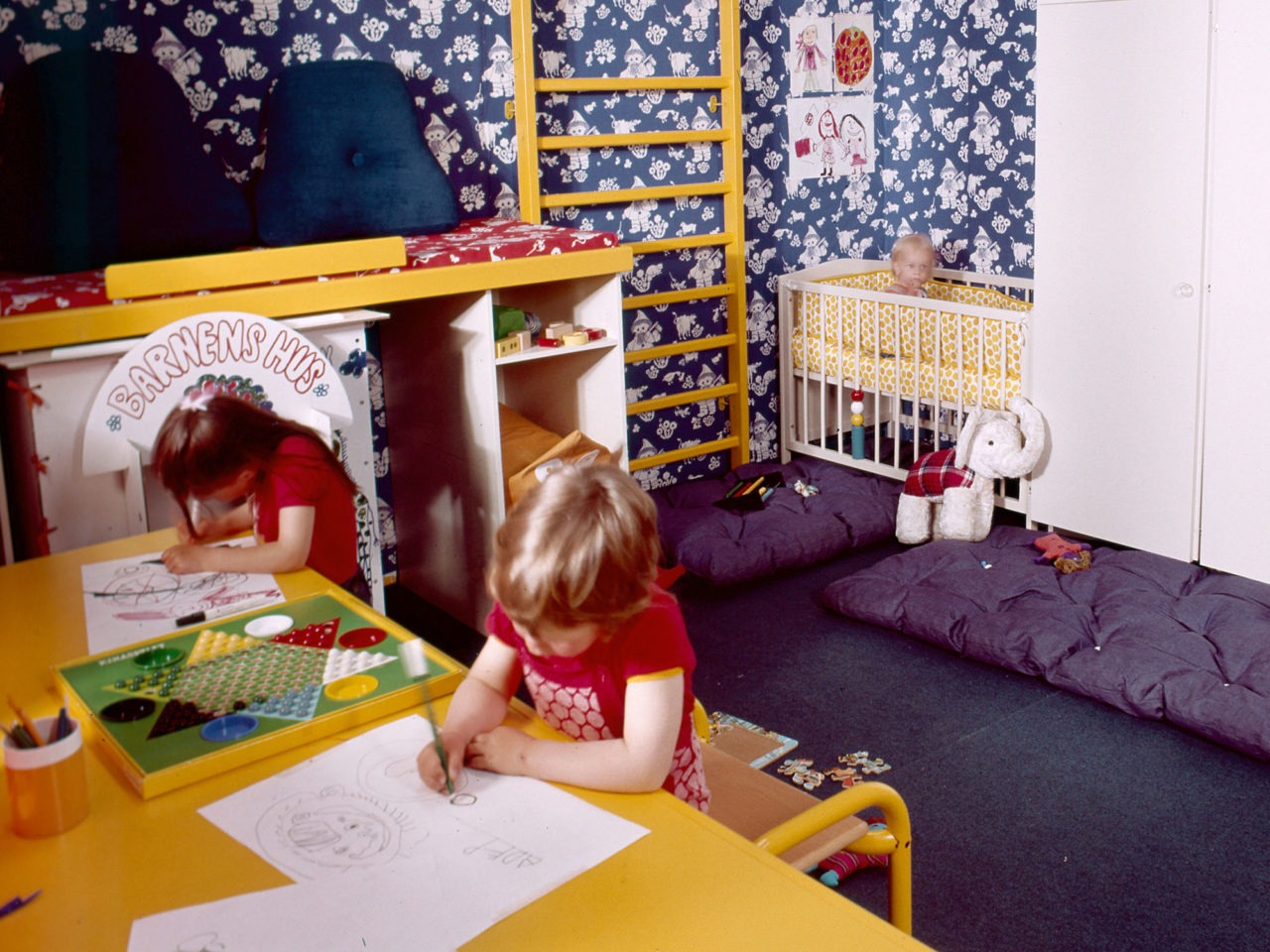 Kinderzimmer. Zwei Kinder zeichnen an langem, gelbem Schreibtisch. Gelbe Sprossenwand an einer Wand mit Sandmann-Tapete.