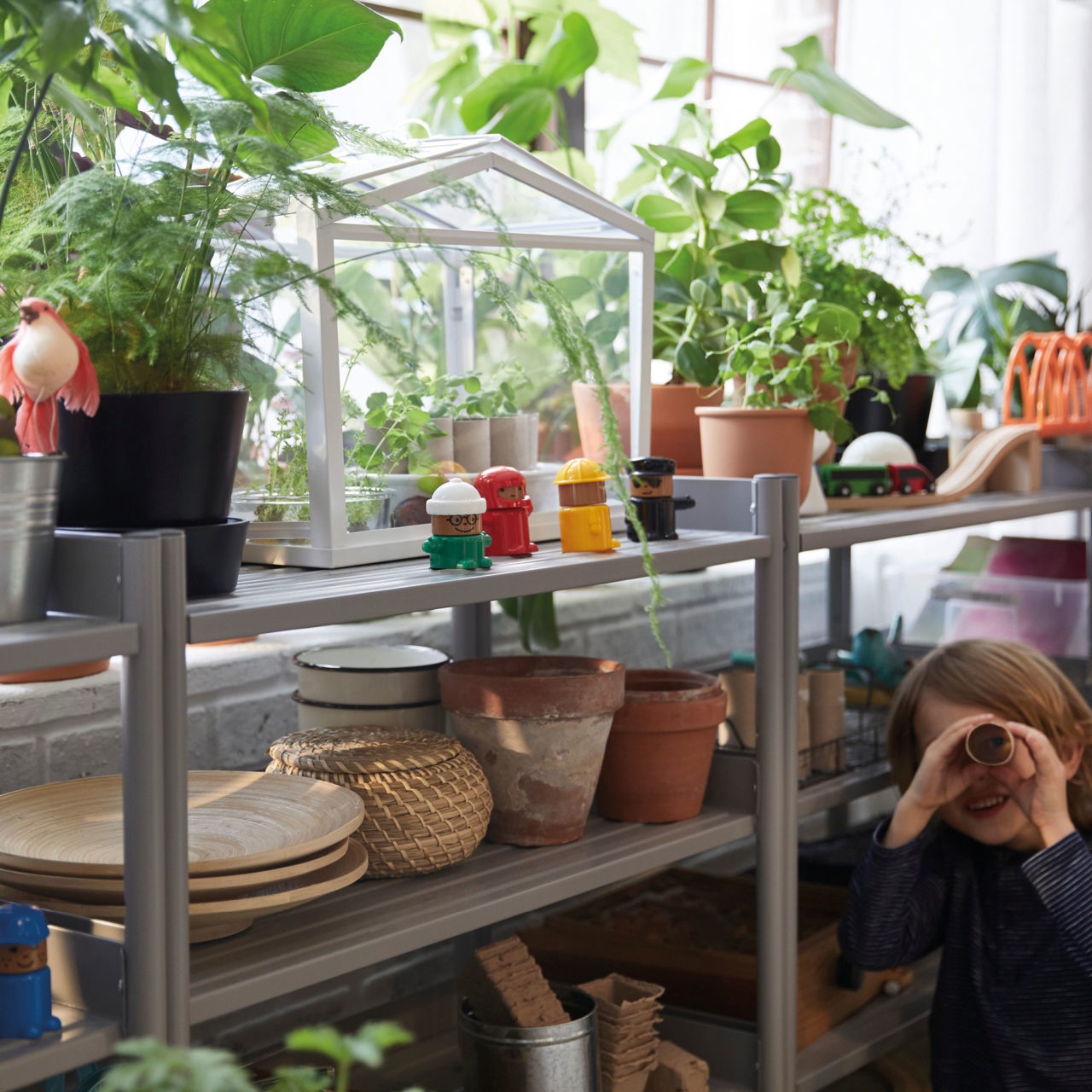 Next to shelves filled with verdant plants and planting accessories, a child stands looking through a paper roll.