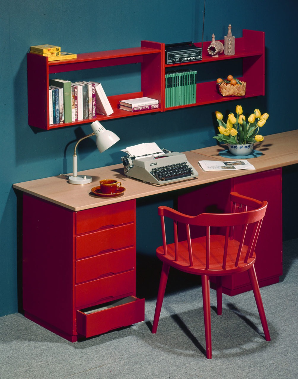 Schreibtisch mit Tischplatte aus hellem Holz und weiteren Details – Wandregale, Schubladenelement und Sprossenstuhl – in Rot.