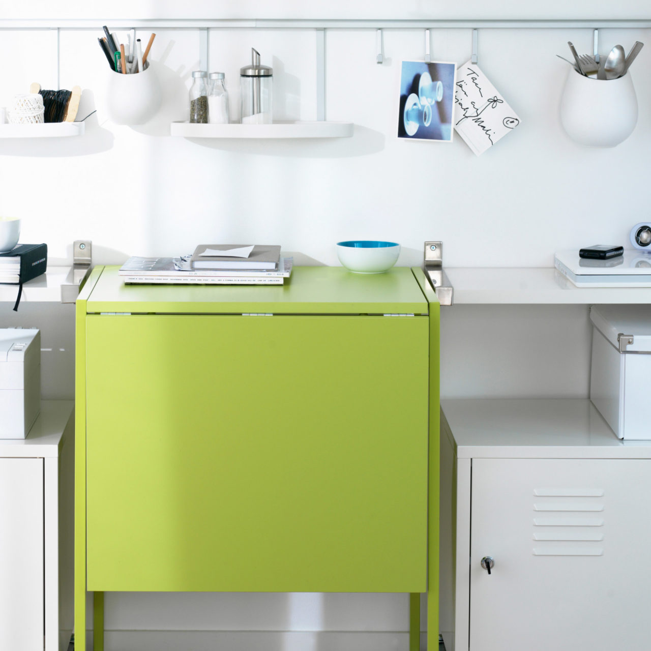 Vit väggsektion där ett limegrönt, nedfällt klaffbord omges av hyllor, metallskåp och småförvaring i vitt.