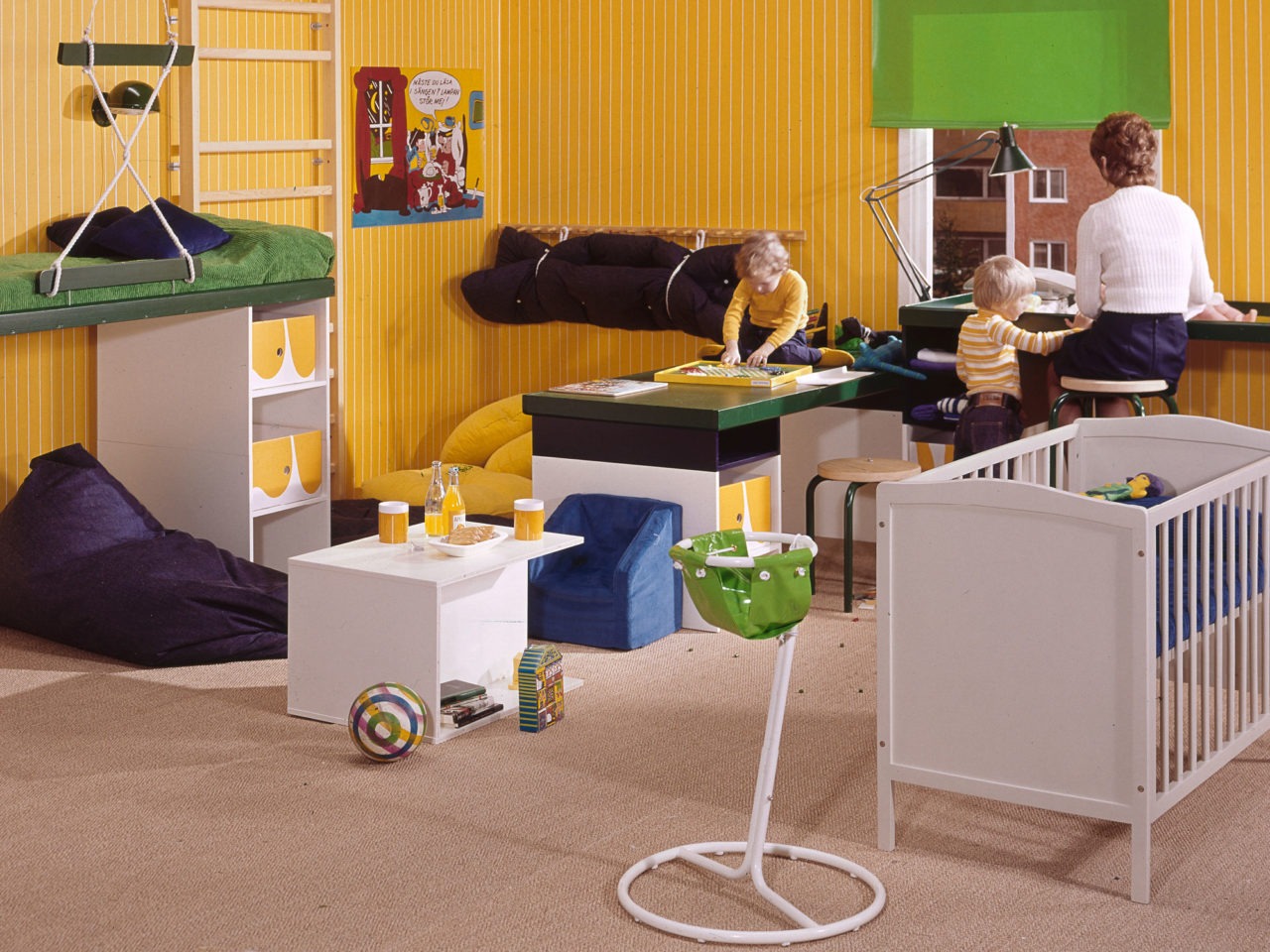 Una mujer sentada frente a un cambiador con un bebé en un cuarto amarillo con una cuna, juguetes y dos niños jugando cerca.