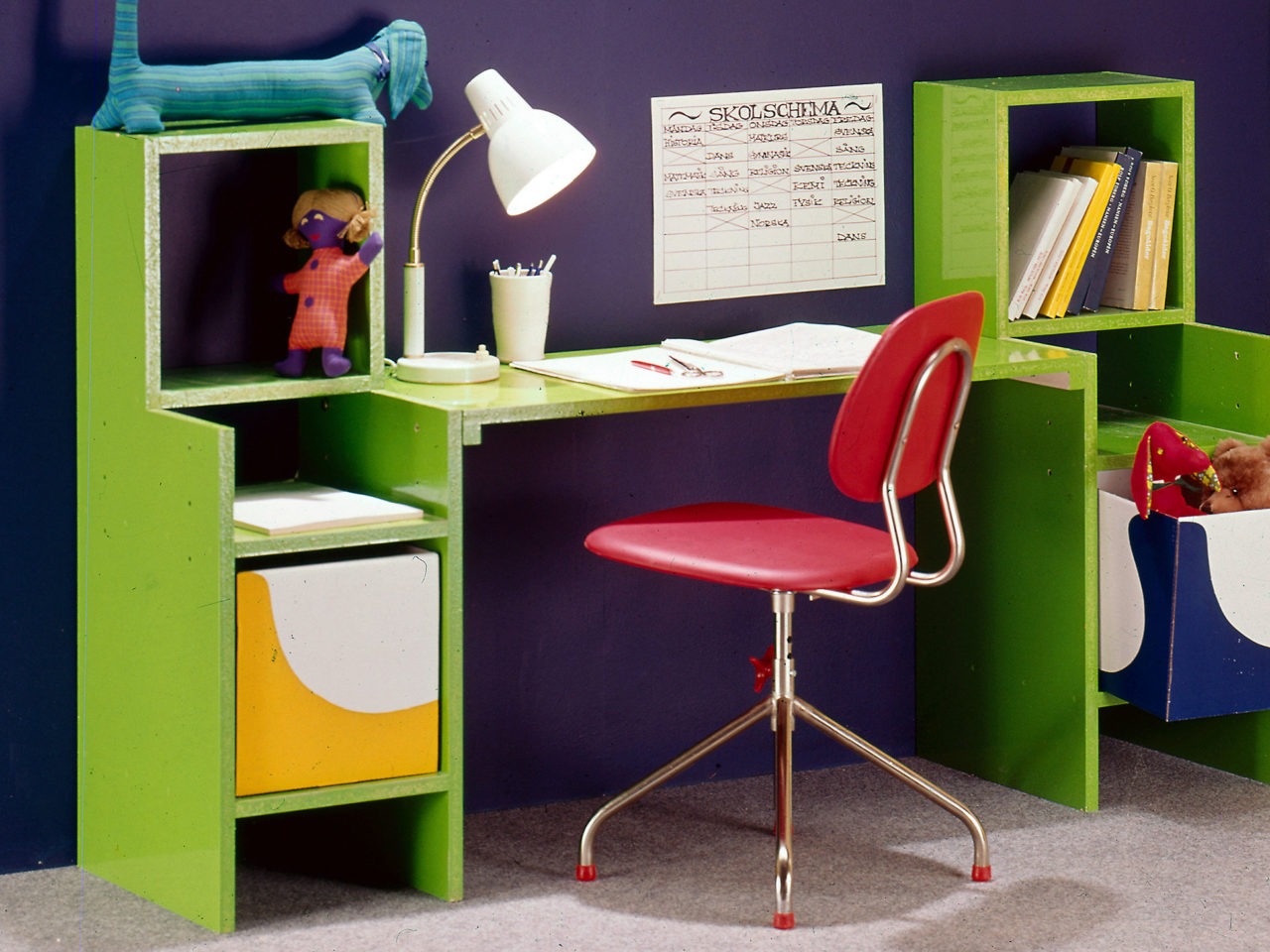 En röd snurrstol står vid en ljusgrön kombination av barnskrivbord och förvaringsmöbel. Mörkblå vägg, grå nålfiltsmatta.