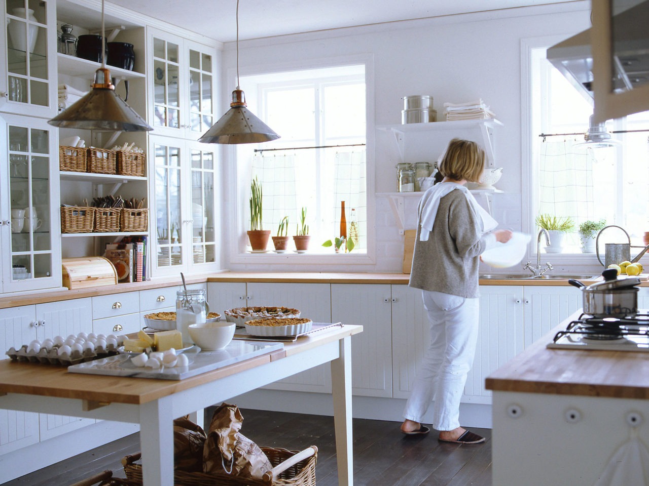 Küche mit weißen Schränken und Arbeitsfläche aus hellem Holz. Eine Frau steht an der Spüle, auf dem Esstisch stehen Kuchen.