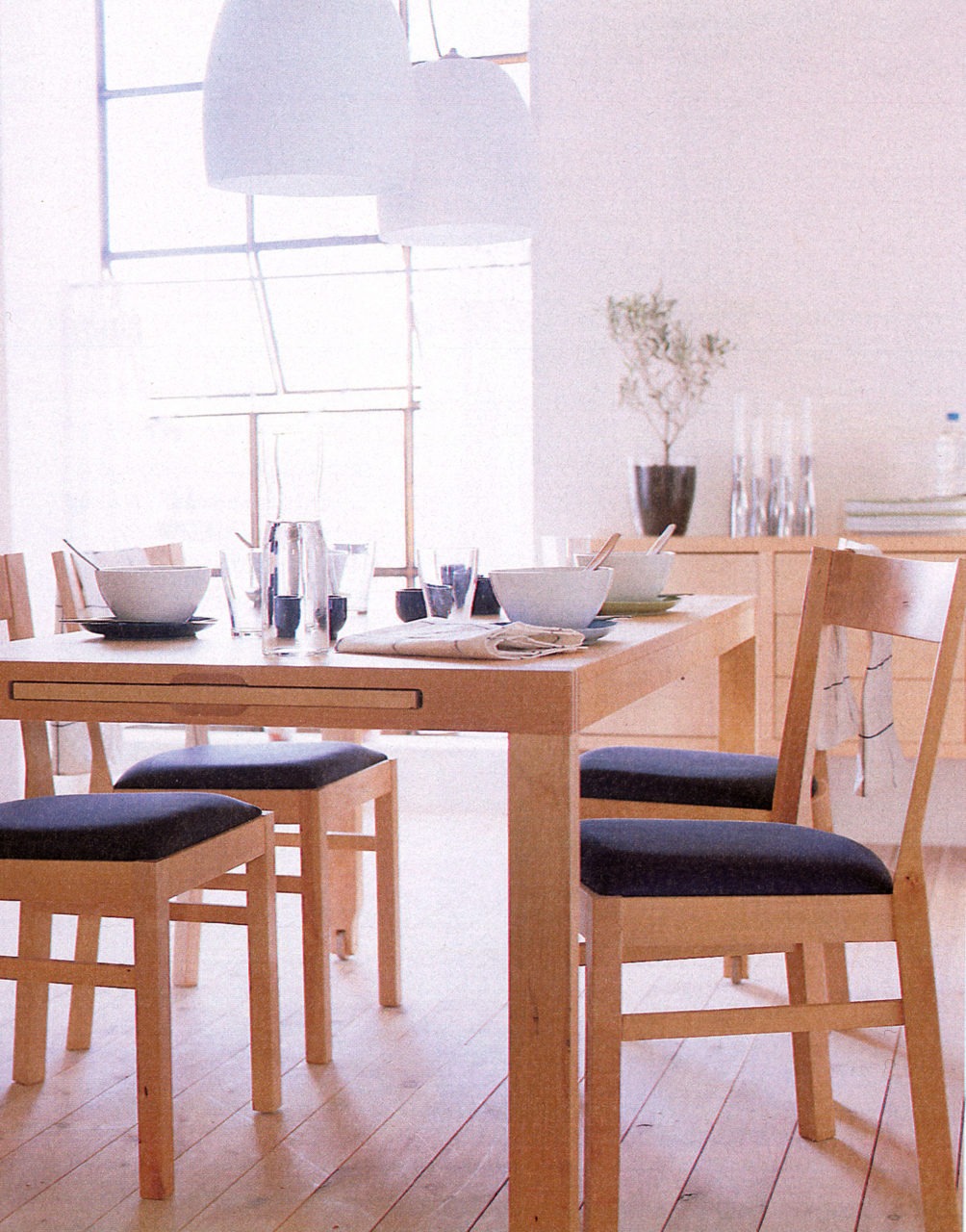 Helle Küche mit Möbeln und hellem Holzfußboden. Stühle mit dunklen Sitzflächen um einen ausziehbaren Esstisch.