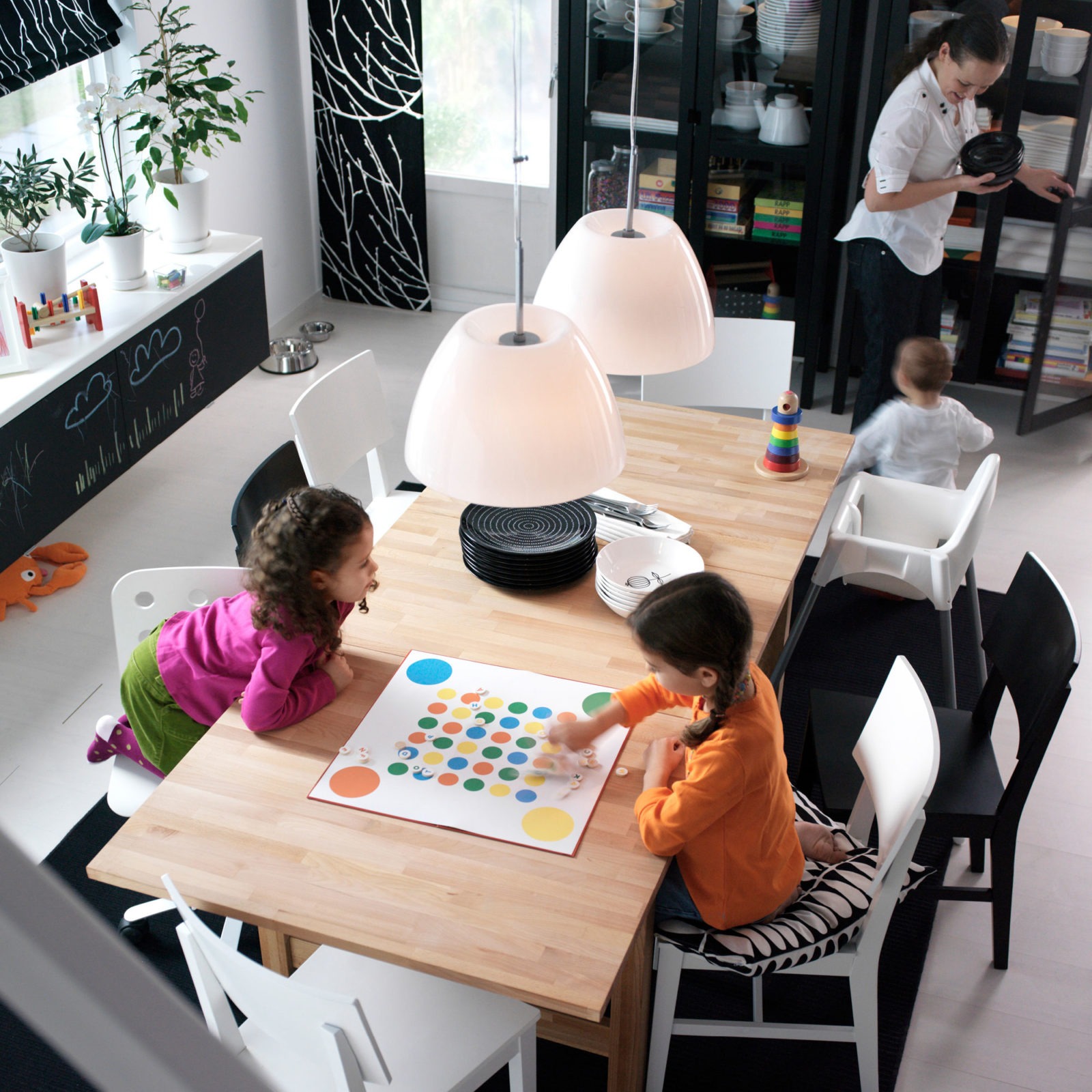 Frau und drei Kinder in einem Raum mit Esstisch aus hellem Holz. Schwarz-weißes Farbkonzept. Zwei Kinder spielen Brettspiel.